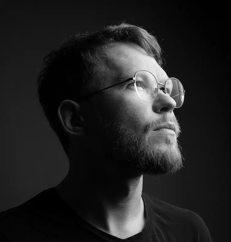 Monochrome          portrait of Andrew Tropin with a neat beard, black t-shirt          and round glasses.  His head is slightly turned to the left.          He is staring into the void and lighting creates a dramatic          contrast.
